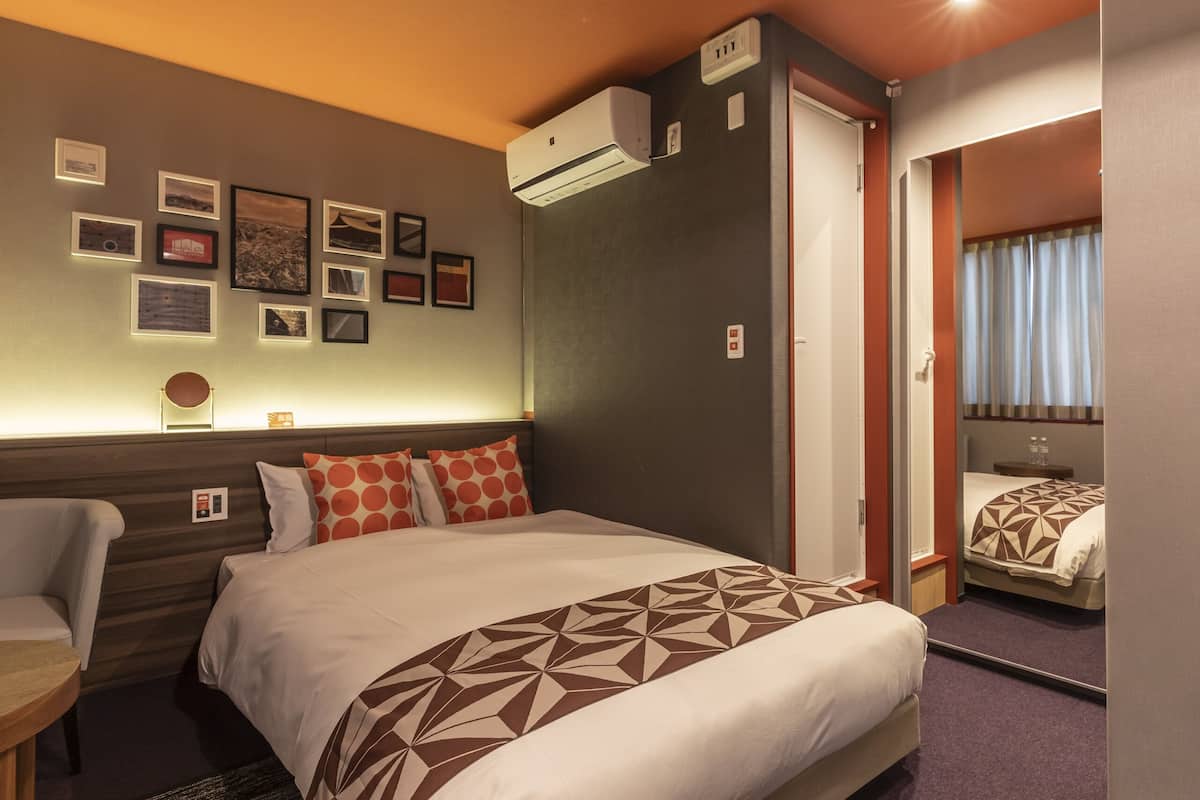 A hotel room with a bed, a chair, a bedside table, a wall-mounted air conditioner, and framed pictures on the wall.