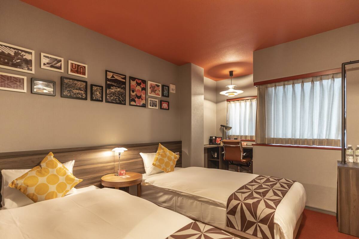 A hotel room with two beds, a desk, a chair, a lamp, and a window with curtains.