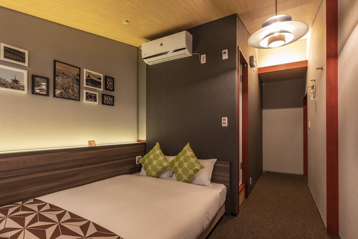 A hotel room with a bed, wall-mounted air conditioning, framed pictures, and a hallway with a door.