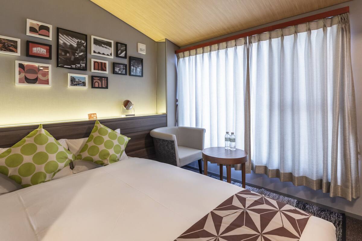 A hotel room with a bed, a small table, a chair, and a window with curtains.