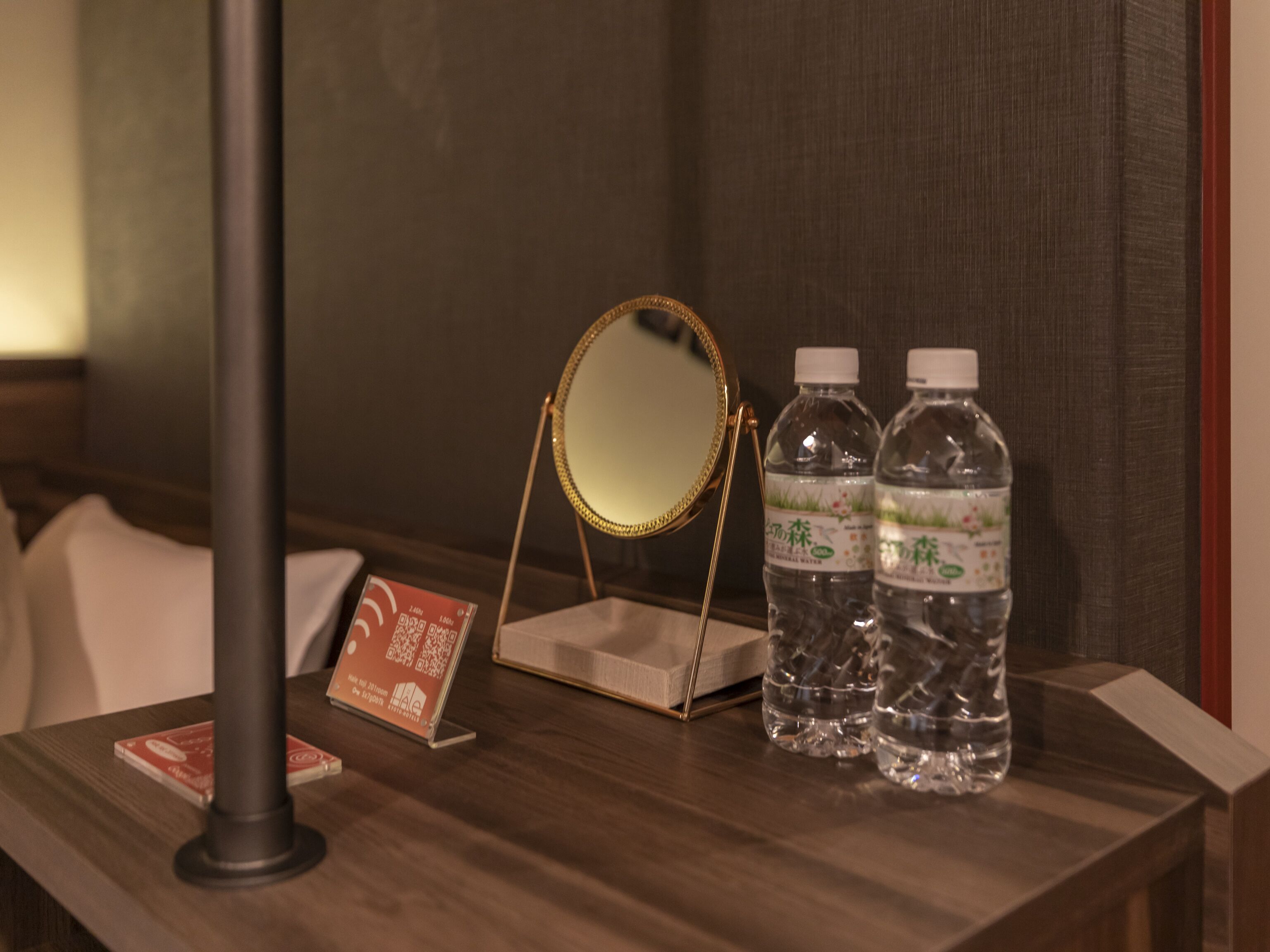 A wooden nightstand with a round mirror, two water bottles, and a QR code stand.