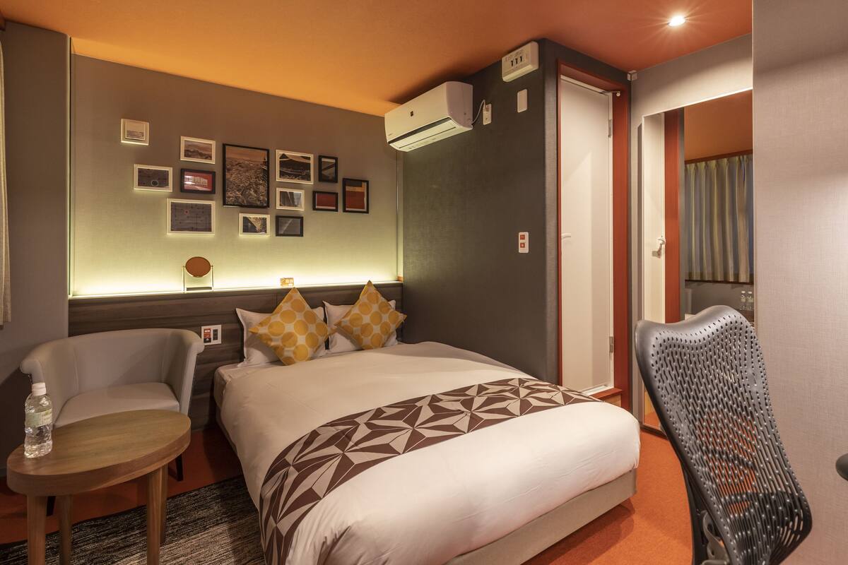 A hotel room with a bed, a chair, a small table, and a wall with framed pictures.