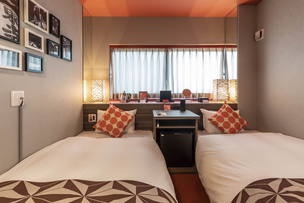 A hotel room with two beds, a desk, a chair, a small fridge, and a window with curtains.