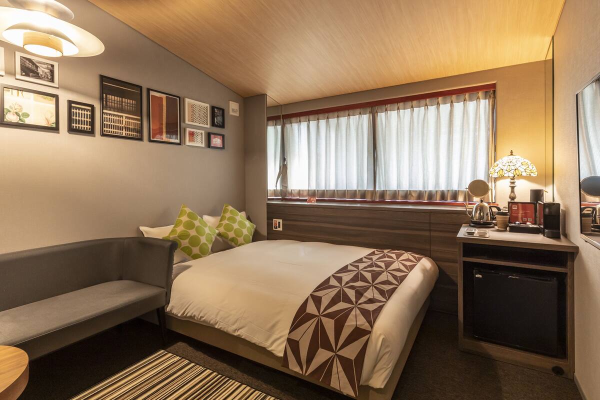 A hotel room with a bed, a desk with a lamp, a small refrigerator, and a window with curtains.