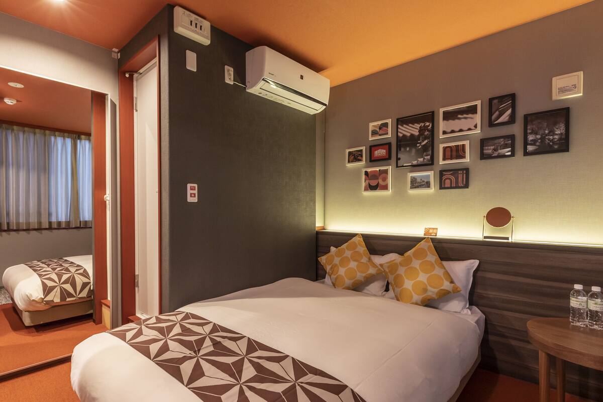 A hotel room with a large bed, a bedside table with water bottles, a wall-mounted air conditioner, and framed pictures on the wall.