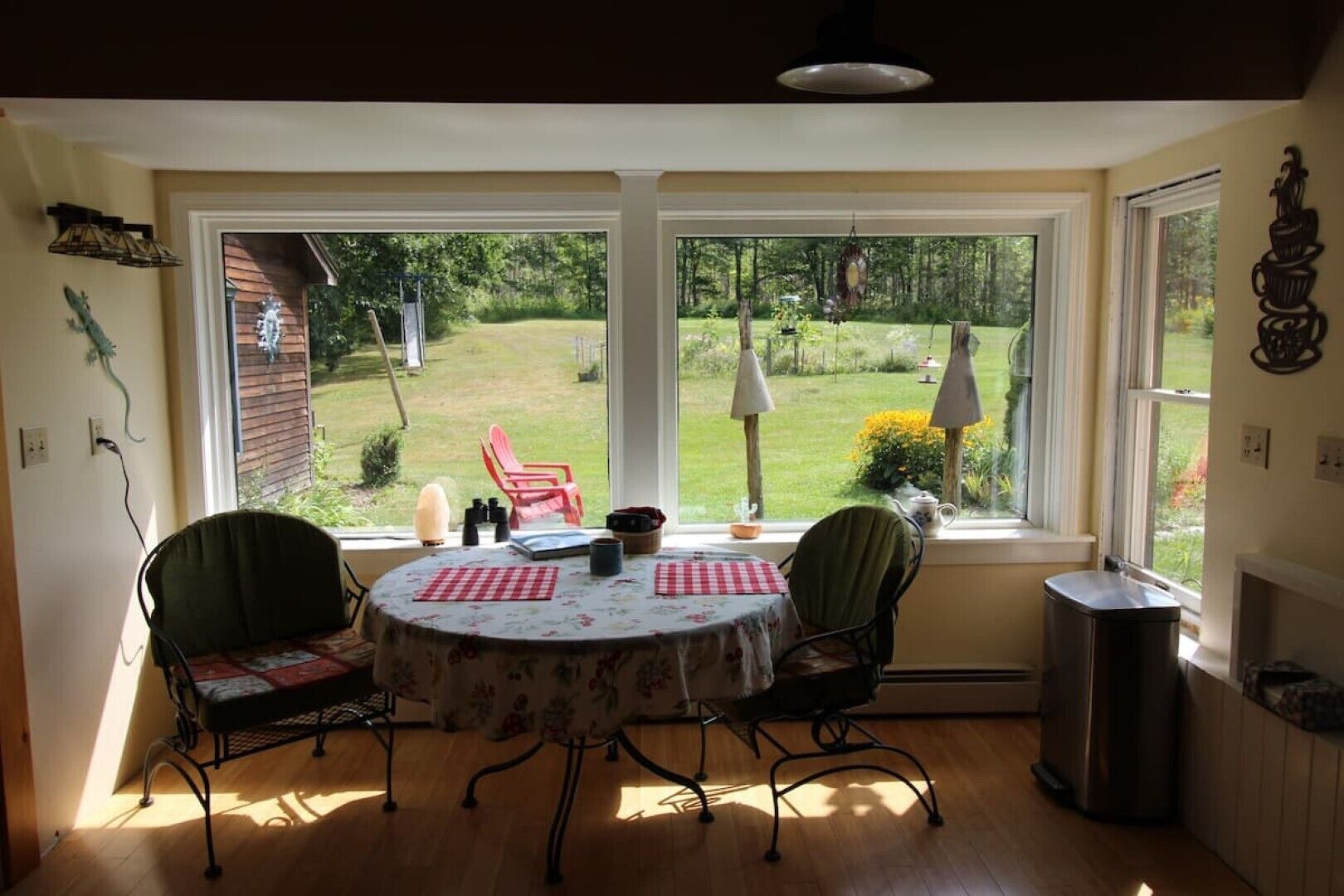 Un coin repas avec une table et des chaises, de grandes fenêtres donnant sur un jardin et une poubelle.