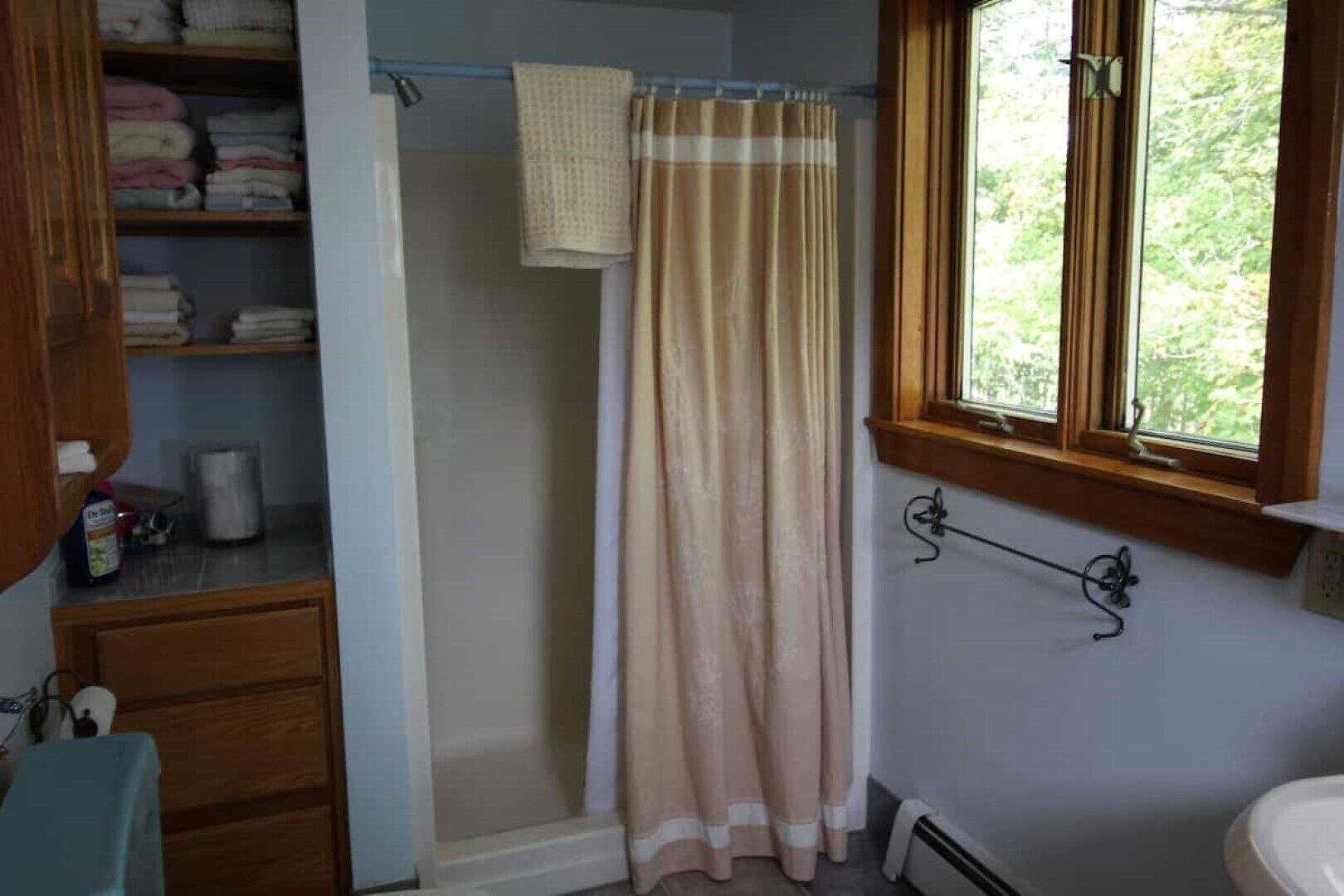 A bathroom with a shower curtain, a towel rack, a window with a view of greenery, and a shelf with folded towels.