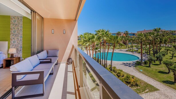 Ein Balkon mit einer Couch, einem kleinen Tisch und einer Lampe. Im Hintergrund ist ein Pool mit Palmen und einem Gebäude zu sehen.