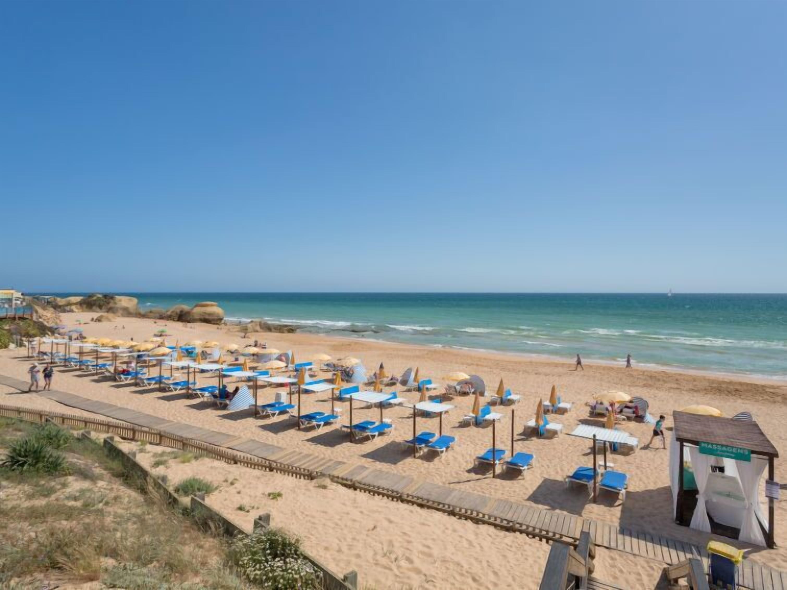 A beach with rows of sun loungers and umbrellas, a wooden boardwalk, and a massage tent.
