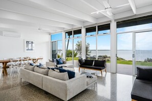 Ein modernes Wohnzimmer mit einer großen Couch, einem Esstisch und Stühlen. Große Fenster bieten Meerblick.