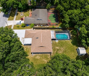 An aerial view of a residential area with a house featuring a large roof, a swimming pool, and a tennis court.