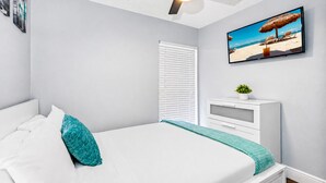 Un dormitorio con cama, ventilador de techo, ventana con persianas, mesita de noche con una planta y un cuadro de temática playera en la pared.