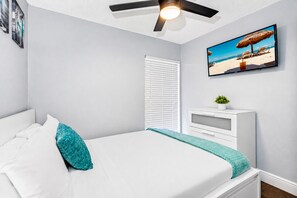 A bedroom with a bed, a ceiling fan, a window with blinds, a bedside table with a plant, and a beach-themed picture on the wall. - Screened Heated Pool, Sleeps 12 | Game Room, 10 Min to Hard Rock Casino (Seffner)