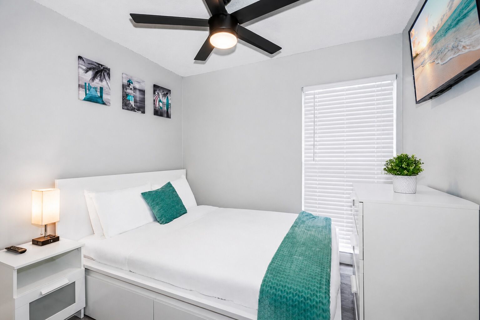 Un dormitorio con cama, mesita de noche, lámpara, ventilador de techo, ventana con persianas, televisor montado en la pared y una pequeña planta en maceta.