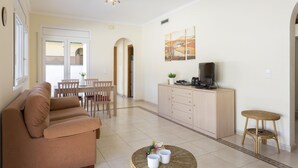 Ein Wohnzimmer mit einer Couch, einem Esstisch mit Stühlen, einer Küchenzeile mit Schrank und einem Fernseher sowie einem kleinen Hocker.