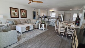A spacious living area with a sofa, coffee table, and dining chairs. A kitchen with white cabinets and a refrigerator is visible in the background. - Cute Bungalow w/small pool!
Absolutely Beach Front! (Gulf Shores)