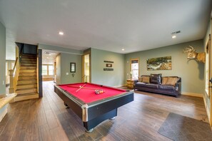 A pool table in a room with wooden flooring, a staircase, a sofa, and a mounted deer head.