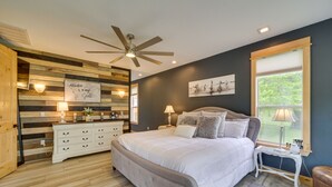 A bedroom with a bed, bedside tables, a ceiling fan, a window with a view, and a wall with a wooden panel design. - Hot Tub, Game Room & More! Lakefront Home in Afton (Afton)