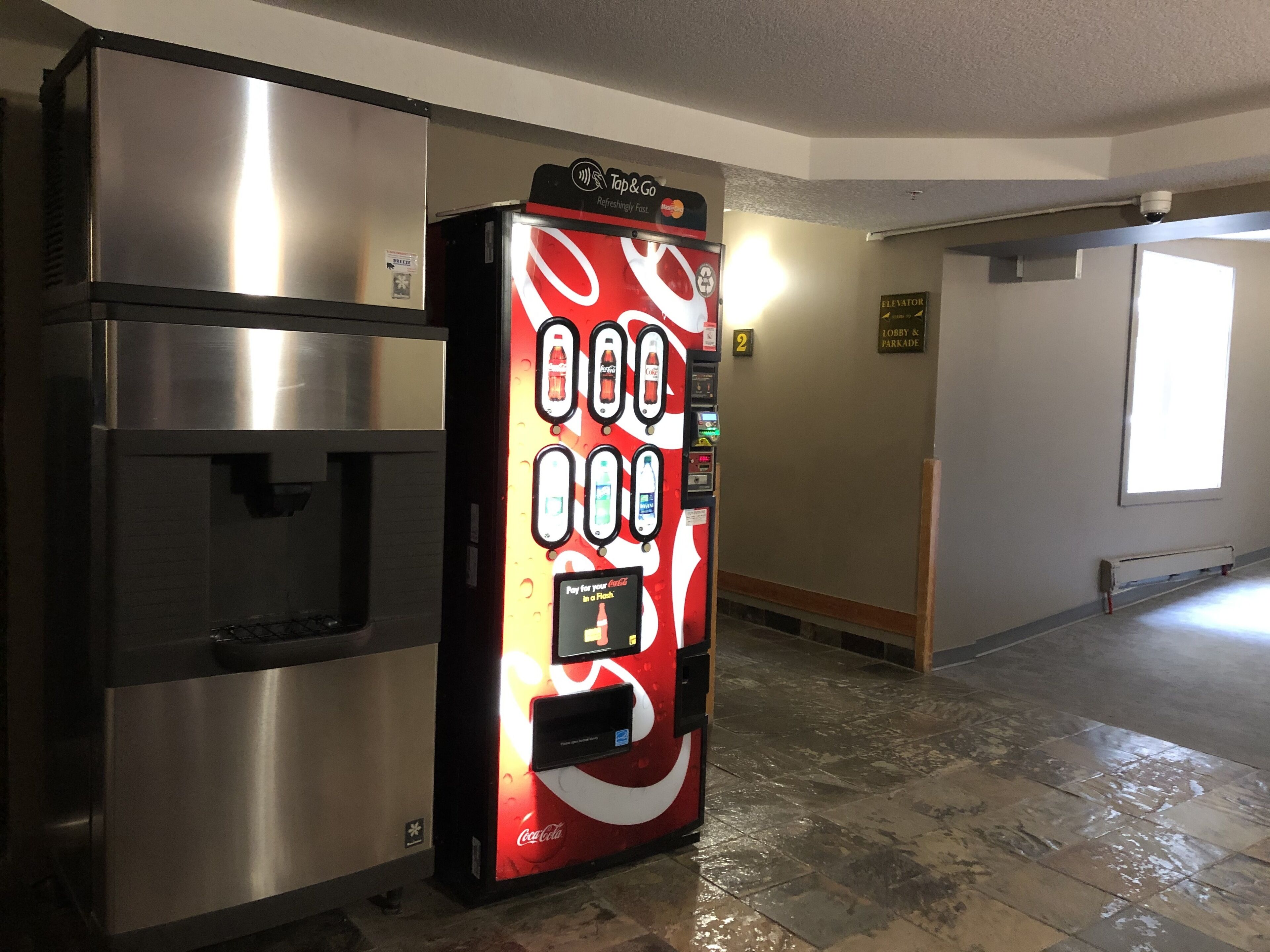 Un distributeur automatique orné du logo Coca-Cola, un réfrigérateur et un panneau indiquant que l’ascenseur se trouve à droite.
