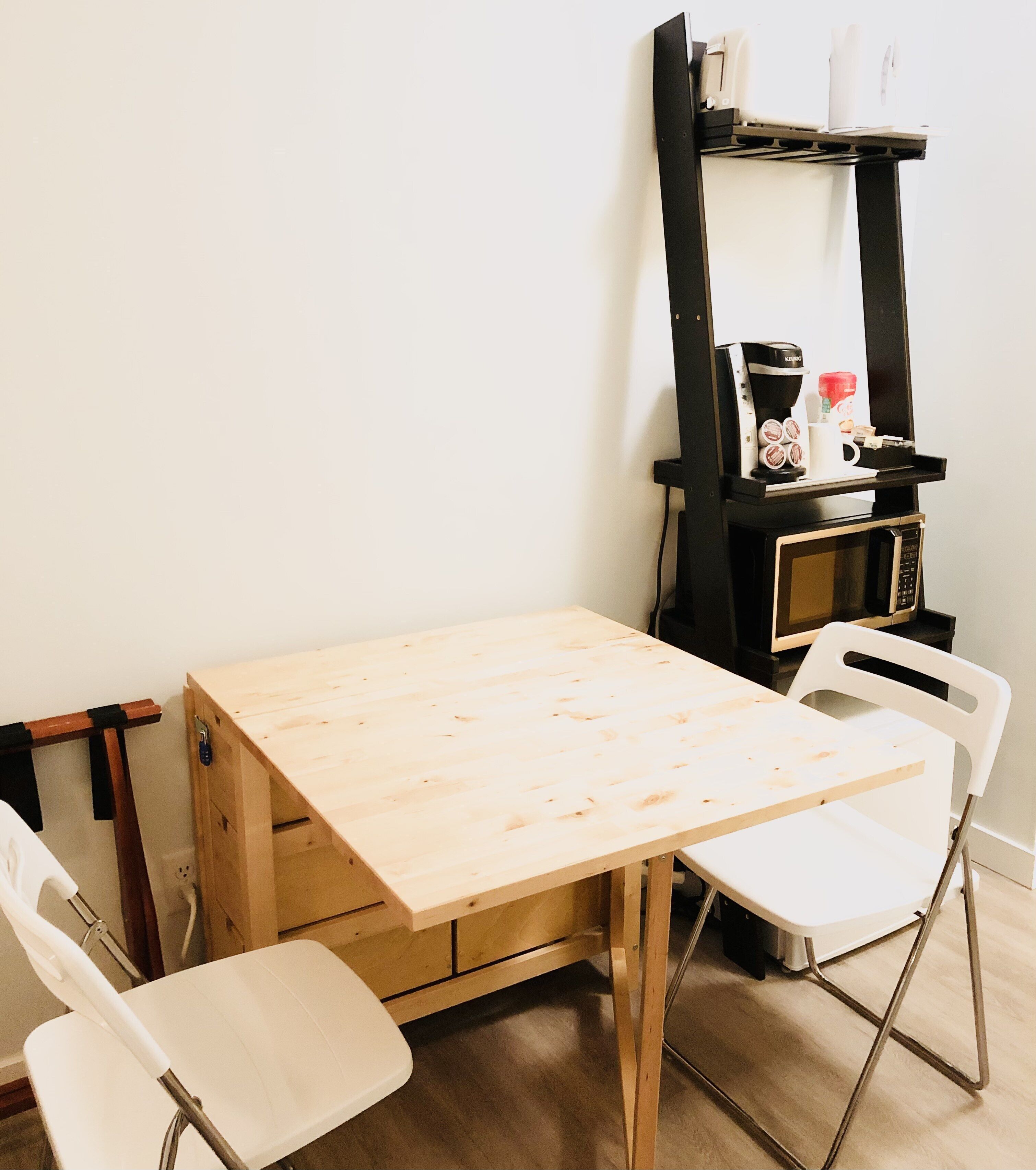 Une petite table en bois, deux chaises blanches, une étagère noire avec une machine à café et un four à micro-ondes.
