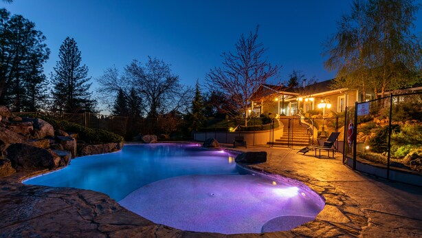A swimming pool with a stone deck, surrounded by a fence and trees. There is a building with lights and a staircase leading to a deck.