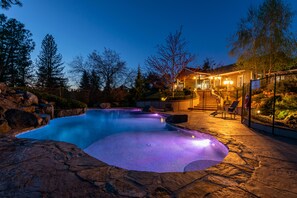 Ein Schwimmbecken mit einer Steindeckung, umgeben von einem Zaun und Bäumen. Im Hintergrund ein Gebäude mit Beleuchtung und einer Treppe, die zu einer Terrasse führt.