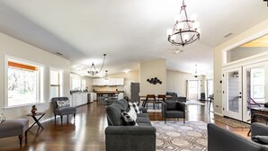 A spacious living area with a gray sofa, a coffee table, and a patterned rug. There is a kitchen in the background with white cabinets and a dining area with a table and chairs.