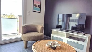 Un soggiorno moderno con una grande TV a schermo piatto, una poltrona confortevole, un tavolino in vimini con oggetti decorativi e vista all'esterno tramite un'ampia finestra.