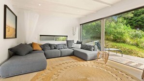 A modern living room with a grey sectional sofa, a woven rug, and a glass door leading to a garden. - Palm Views - 3-Minute walk to Palm Beach (Waiheke Island)