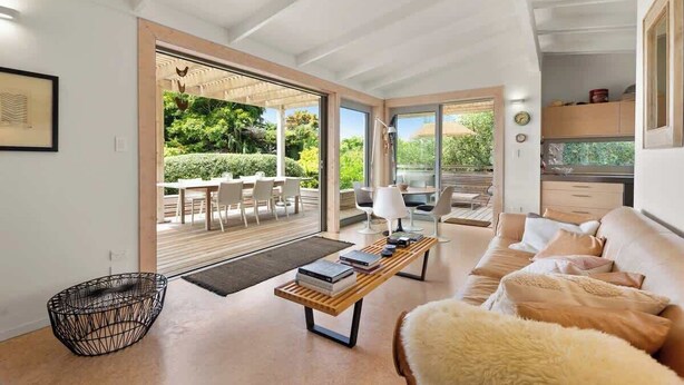 A modern living room with a sofa, a coffee table, and a dining area with chairs and a table. - Palm Views - 3-Minute walk to Palm Beach (Waiheke Island)