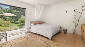 A modern bedroom with a large window, a bed, a bedside table, and a view of a garden. - Palm Views - 3-Minute walk to Palm Beach (Waiheke Island)