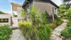 A modern house with a sloped roof, a wooden deck, and a gravel pathway leading to the entrance. - Palm Views - 3-Minute walk to Palm Beach (Waiheke Island)
