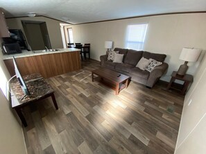 A living room with a sofa, coffee table, and a kitchen area with a counter and chairs. - Custom Touch Village-Sulphur (Sulphur)