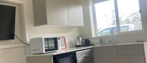 Eine kompakte Küche mit Mikrowelle, Toaster und Geschirrspüler. Ein Fenster sorgt für natürliches Licht, und ein kleiner Mülleimer ist sichtbar.