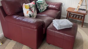 A red leather sofa with two patterned pillows, a matching ottoman, a small wooden side table with a lamp, and a woven rug.