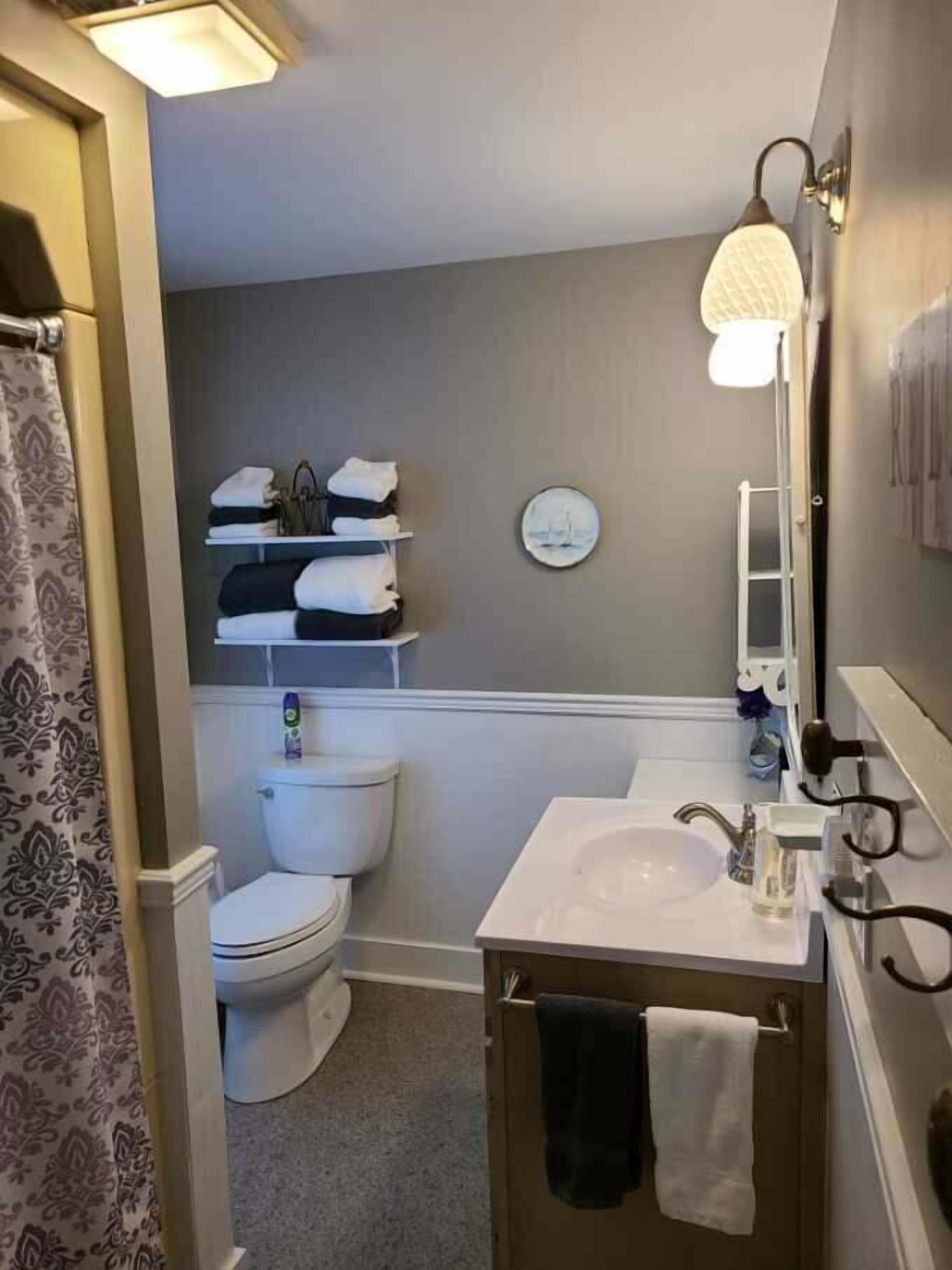 Et badeværelse med toilet, vask og håndklæderække. Der er en hylde med sammenfoldede håndklæder og et rundt spejl over vasken.