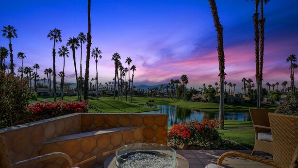 A patio with wicker chairs and a glass-top table, overlooking a golf course and a body of water, with palm trees and red flowers in the foreground. - Access to Gym/Golf/Tennis/Pickleball via platinum membership. (Palm Desert)