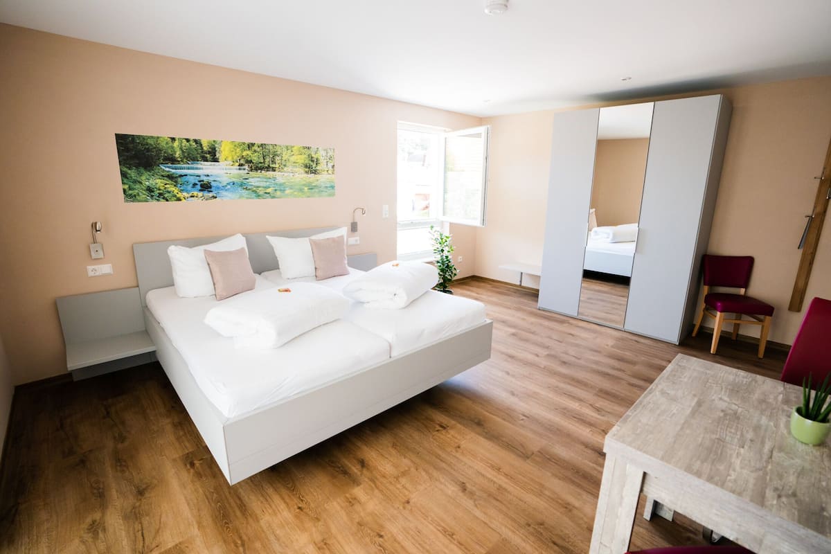 A modern bedroom with a large bed, wooden flooring, a painting on the wall, and a view through an open door.