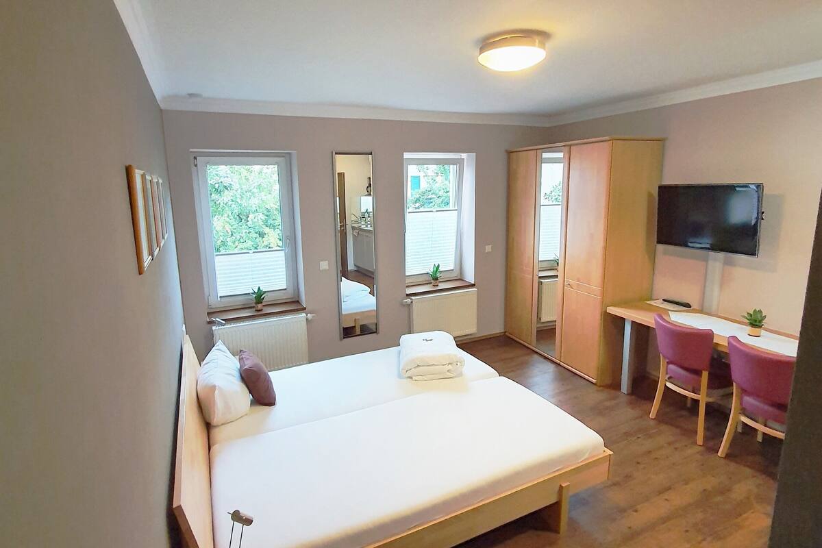 A bedroom with a bed, a desk, a chair, a TV, and a window.
