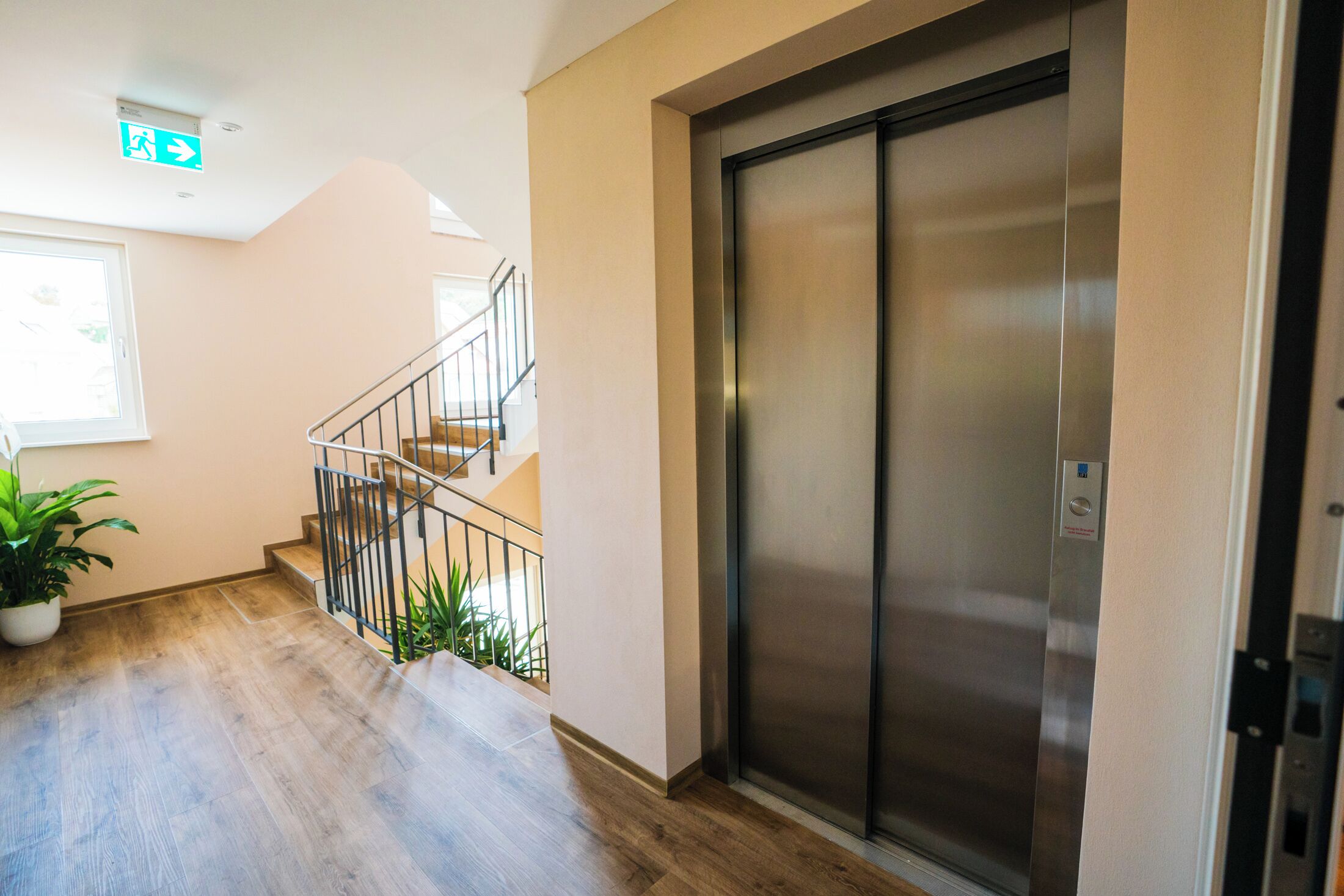 A modern elevator with a metallic finish, a staircase with a metal railing, and a potted plant on the floor.