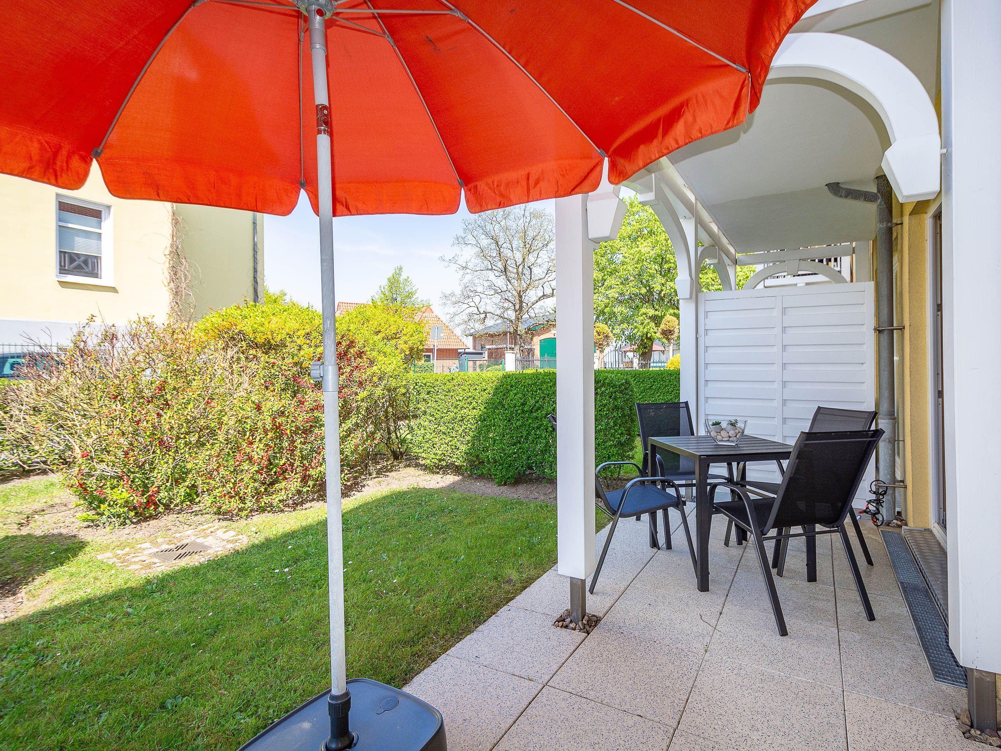 Un patio al aire libre con una mesa, sillas y un paraguas rojo, con un edificio blanco al fondo.