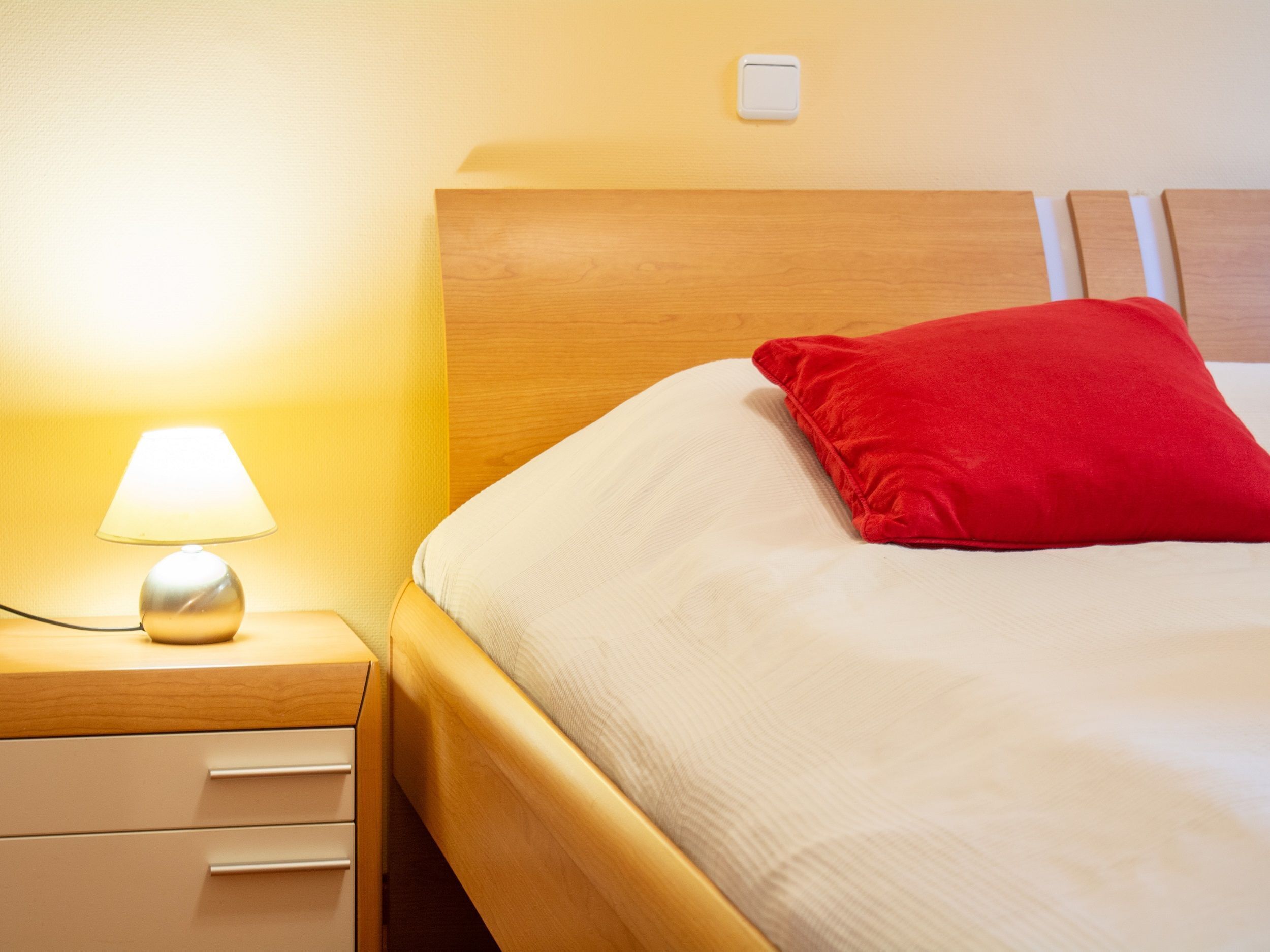 A neatly made bed with a red pillow, a bedside lamp, and a wooden nightstand with drawers.