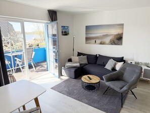 En moderne stue med sofa, lænestol, sofabord og udsigt til naturen gennem en skydedør.