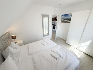 Et soveværelse med seng, et natbord, en lampe, et spejl og udsigt til et køkken og badeværelse gennem en åben dør.