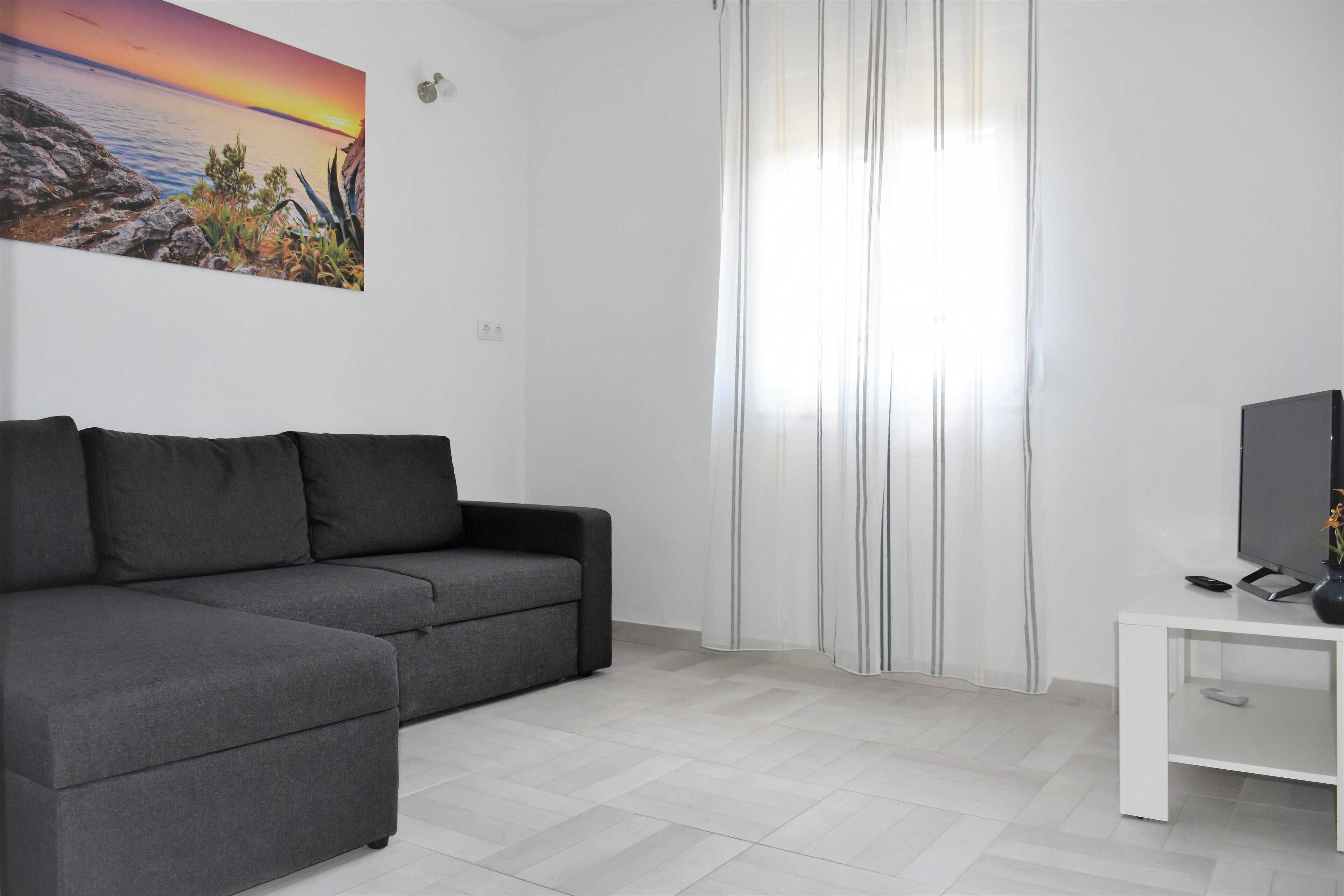 A modern living room with a grey sofa, a flat-screen TV on a white stand, and a painting of a sunset over water on the wall.