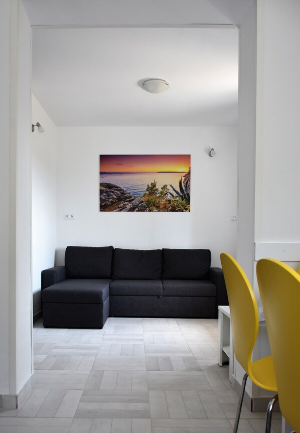 A modern living room with a dark grey sectional sofa, a yellow chair, and a painting of a sunset over a rocky landscape on the wall. - Apartment Meryl (Solin)