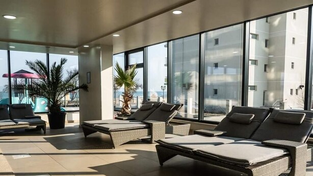 Espaço de sala de estar iluminado pelo sol, com poltronas de vime e uma grande janela com vista para o mar.