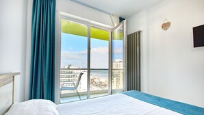 Quarto de hotel com uma janela grande, uma cama, uma poltrona e vista para a praia e edifícios.
