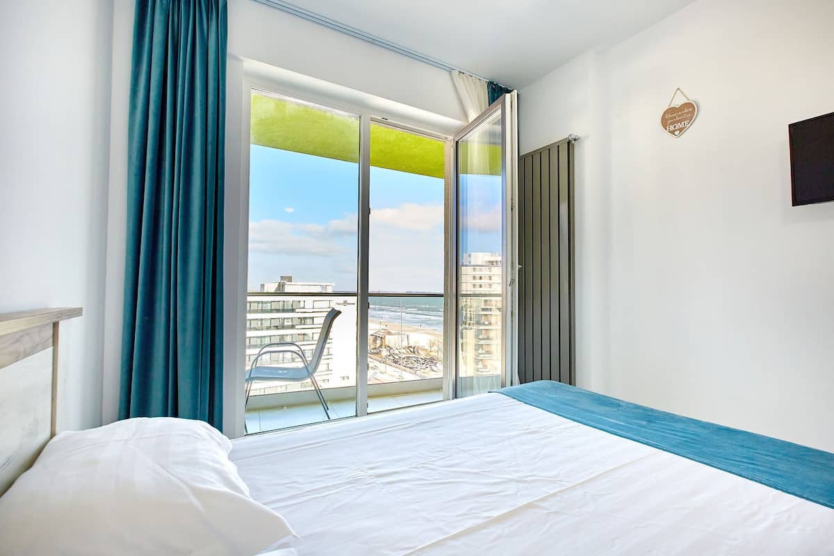 A hotel room with a large window, a bed, a chair, and a view of the beach and buildings.