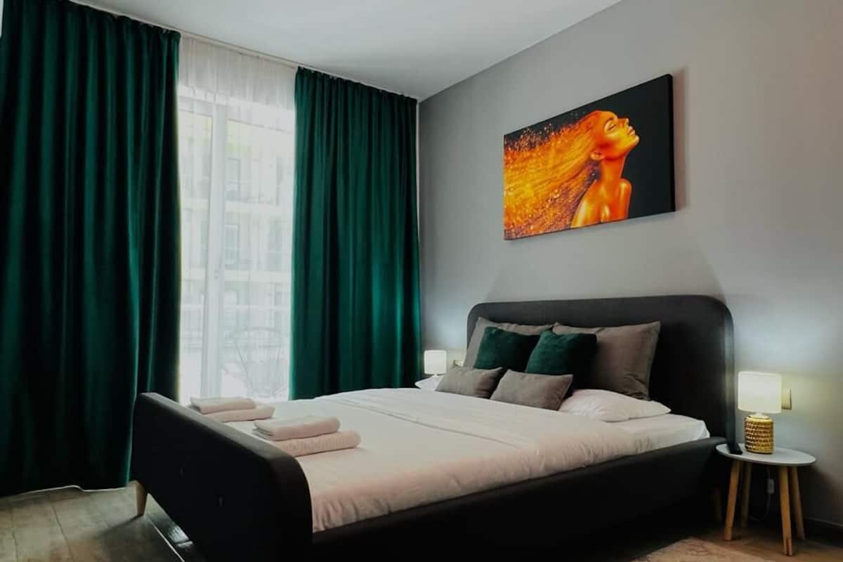 A modern bedroom with a large bed, green curtains, a painting on the wall, and a bedside lamp.